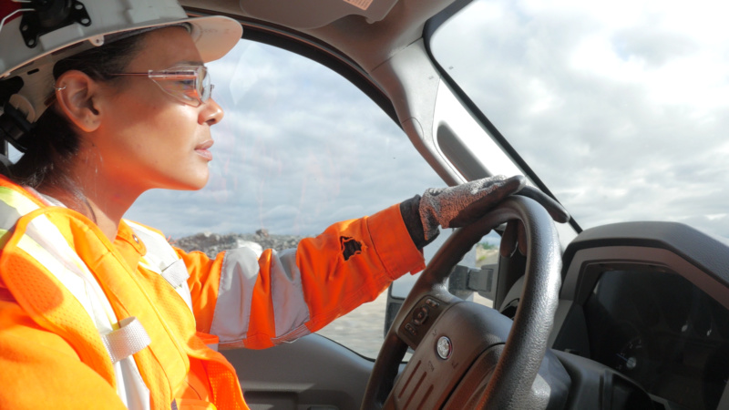 A visible minority woman wearing an orange reflective safety vest and driving a truck