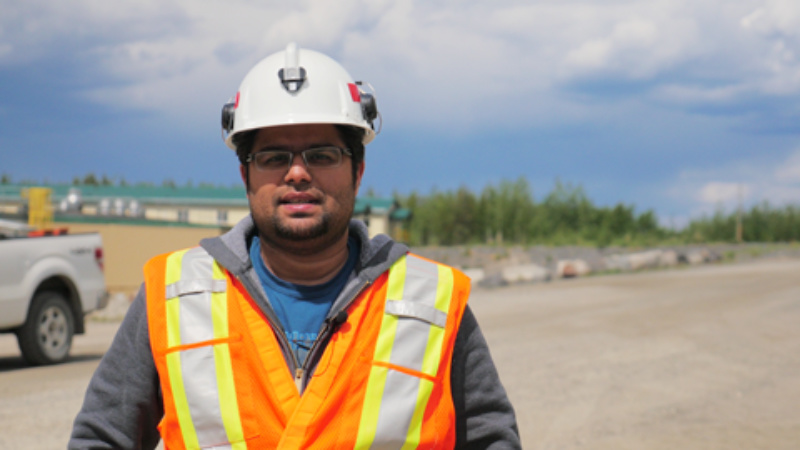 A visible minority man wearing a white hard hat and orange reflective safety vest and standing in front of a road