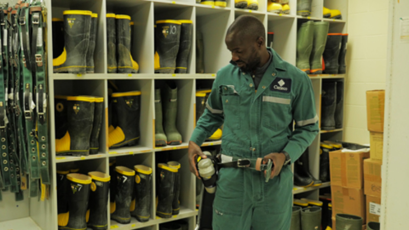 A visible minority male putting on a safety belt in a room that contains storage of personal protective equipment. Safety boots and belts are in the background.